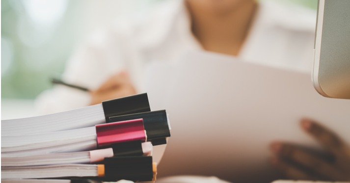 woman reading a document with a pen in hand and there is a pile of other documents on the desk in front of her