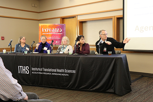 4 women and one man sitting in a row at a table behind microphones. The man, at left is gesturing.