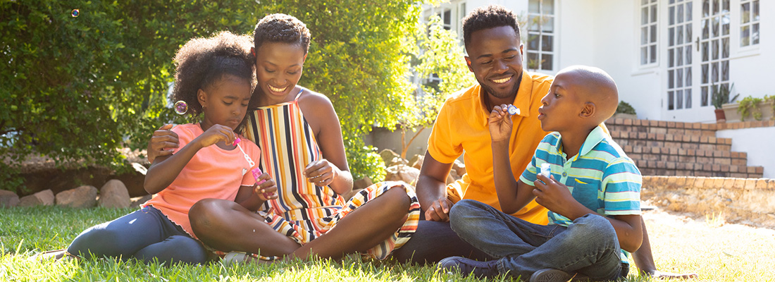 A black family, mother, father, daughter and son, sitting together on their lawn in bright sunshine, blowing bubbles and smiling.