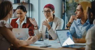 a group of people sitting around a table talking and working on laptops