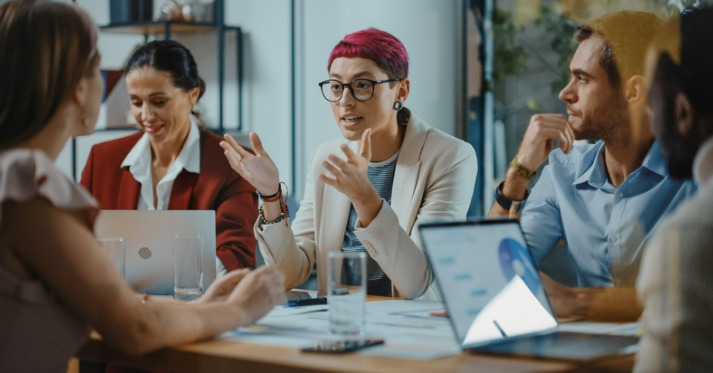 a group of people sitting around a table talking and working on laptops